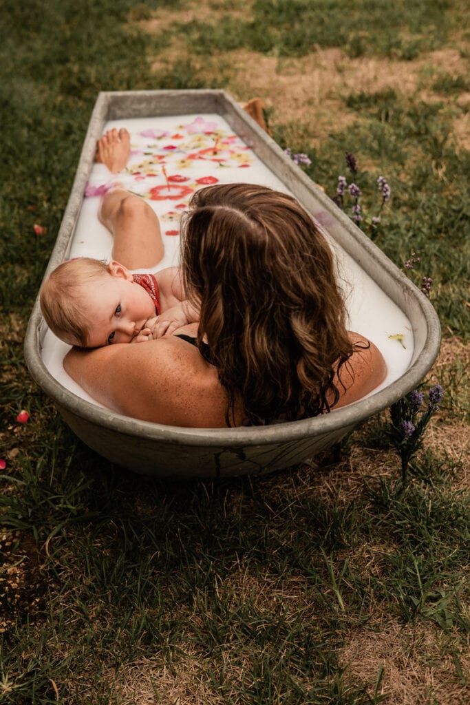 Motherhood Photographer, a mother holds her baby in an outdoor tub filled with milk and flowers
