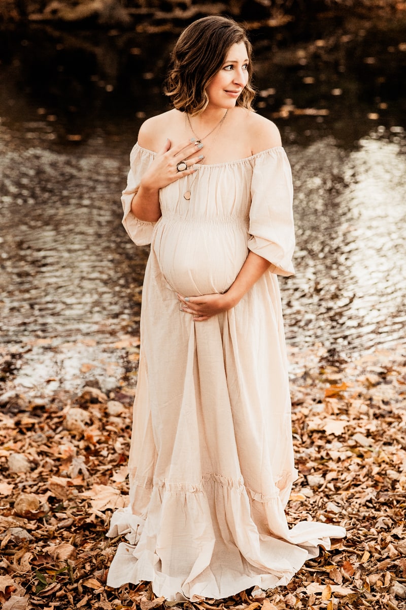 Maternity Photographer, An expectant mother to be holds her pregnant belly. she stands near a quiet river in the fall leaves.