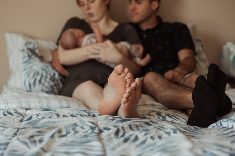 Motherhood Photographer, a new mother and father sit on the bed admiring their newborn baby in mom's arms